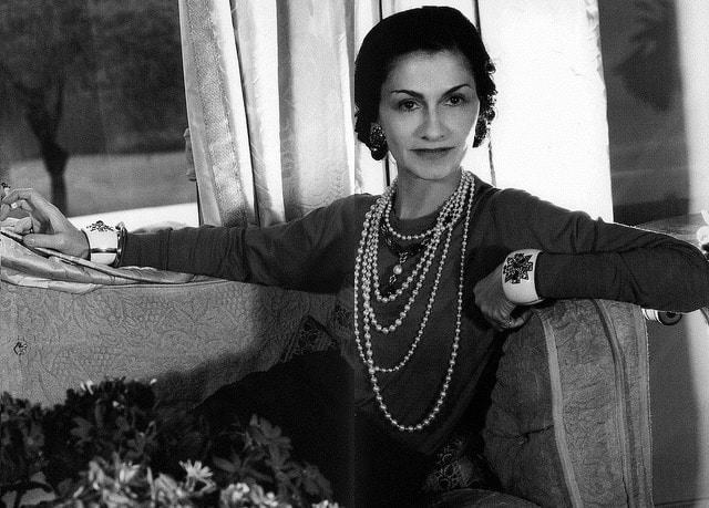 een wit-zwart foto van Coco Chanel waarop te zien is dat ze in de hoek van een zetel zit. Met vijf lange parelkettingen aan, in verschillende lengtes. Ook twee grote witte armbanden en ze kijkt zelfzeker in de camera.